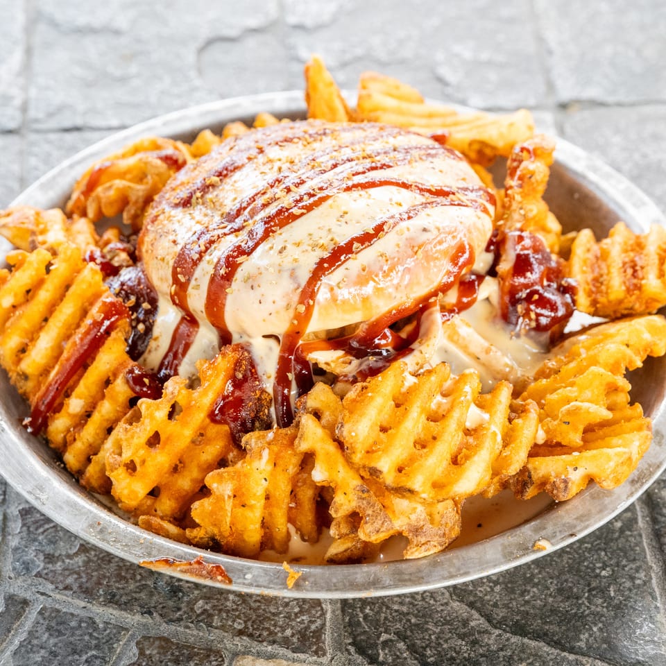 Loaded burger topped with creamy and barbecue sauces, surrounded by curly and waffle fries on a metal plate.