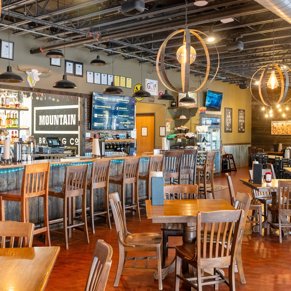 Spacious rustic restaurant interior with wooden tables, bar seating, exposed ceiling, pendant lights, and a stocked liquor bar.