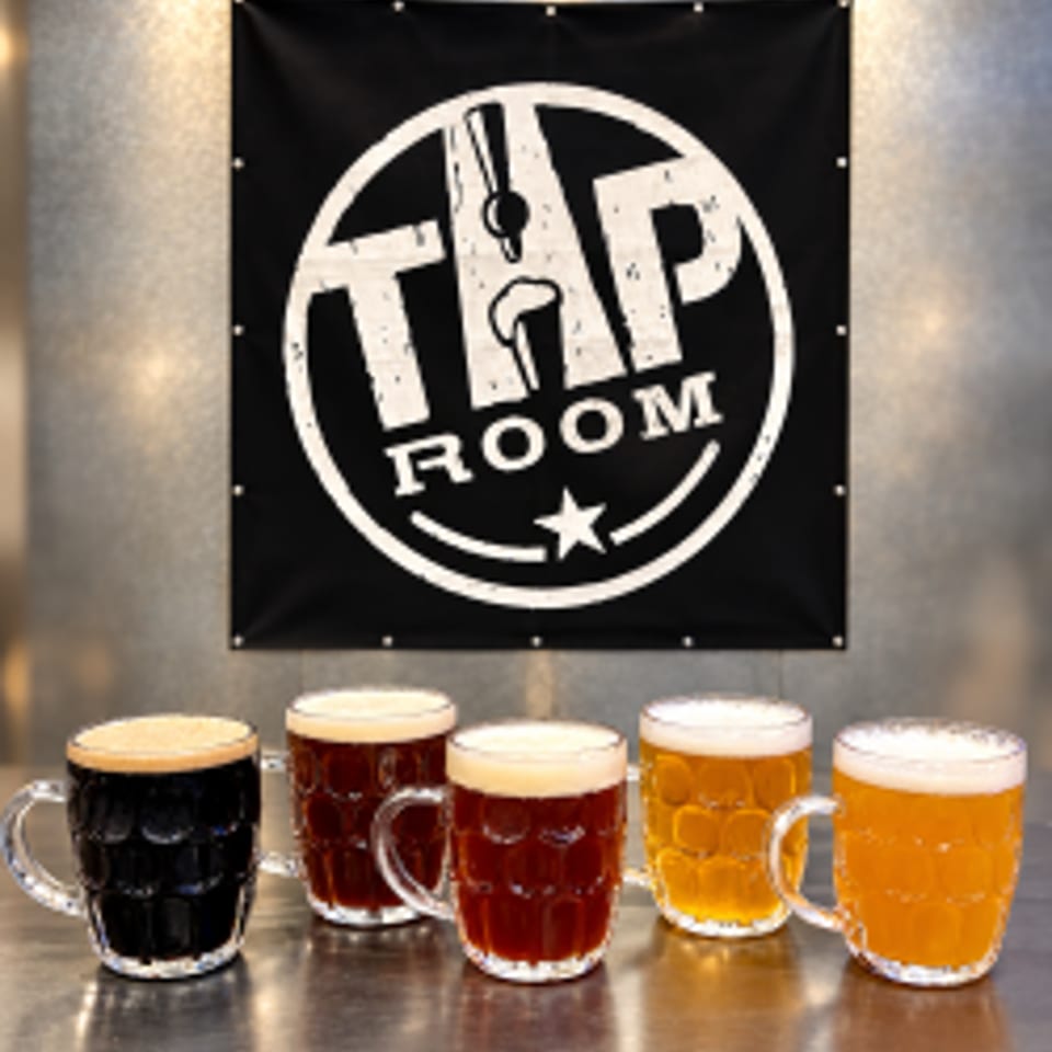 Five beers in tasting glasses sit beneath a large Tap Room sign on a textured wall.