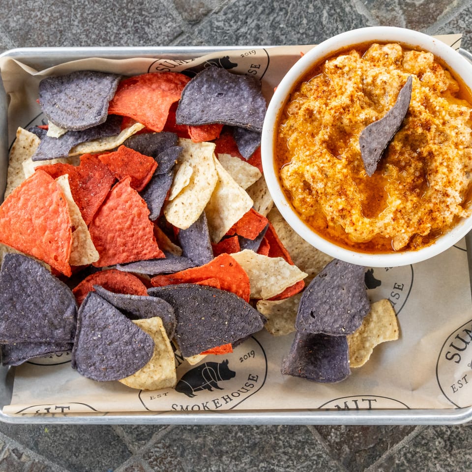 Overhead view of colorful tortilla chips served with a bowl of warm, seasoned cheese dip on a metal tray.