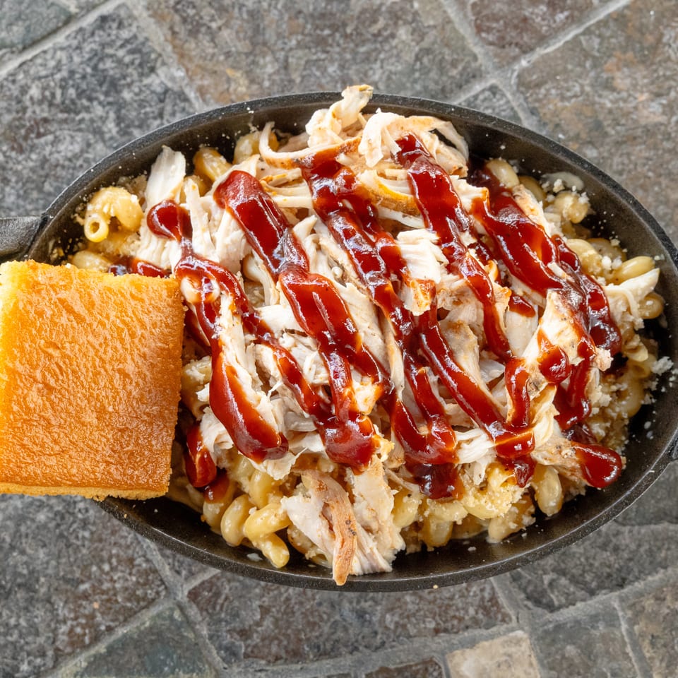 Cast-iron skillet of macaroni and cheese topped with shredded chicken and barbecue sauce, served with cornbread.