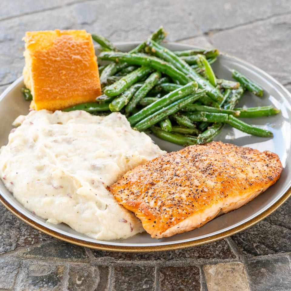 Grilled salmon with mashed potatoes, green beans, and cornbread served on a plate atop a stone table.