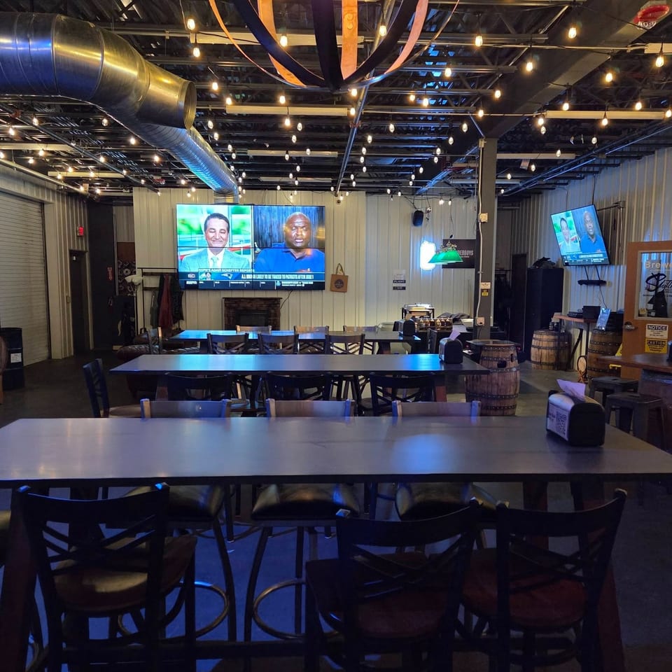 Industrial-style restaurant dining area with long tables, stools, string lights, exposed ductwork, colorful ceiling streamers, and multiple televisions.