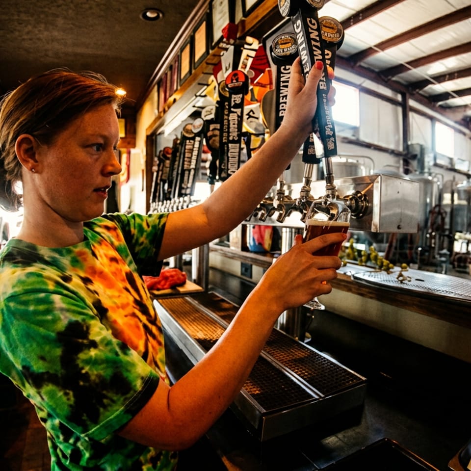 Bartender in a tie-dye shirt pouring a beer from a tap at a brewery bar.