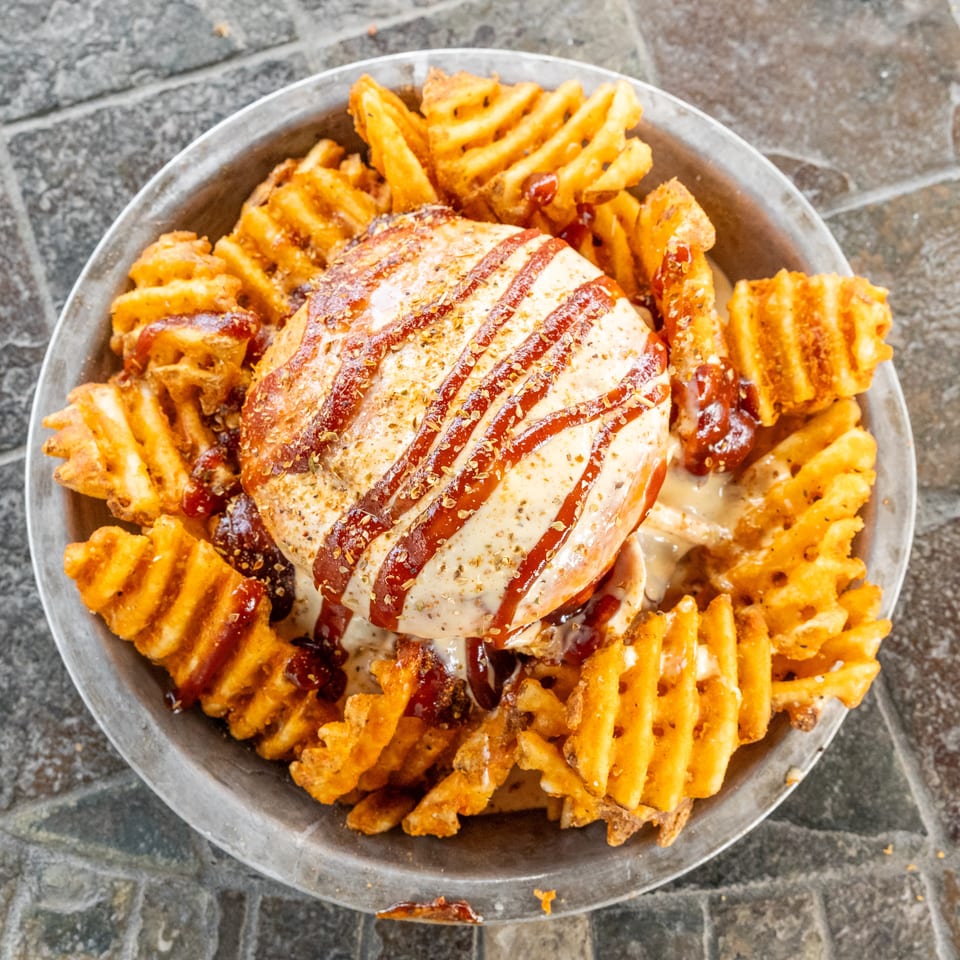 Burger topped with melted cheese, ketchup, and herbs, surrounded by crispy waffle fries on a metal plate.
