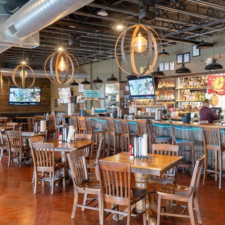 Spacious restaurant dining room with wooden tables, bar, exposed ductwork, decorative pendant lights, televisions, and framed wall art.