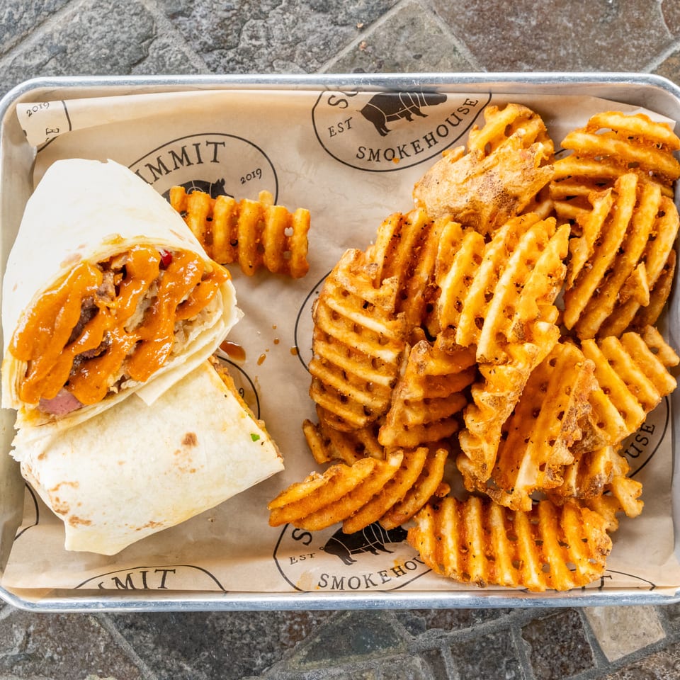 Tray holding a saucy wrap and a large serving of golden waffle fries on branded paper.