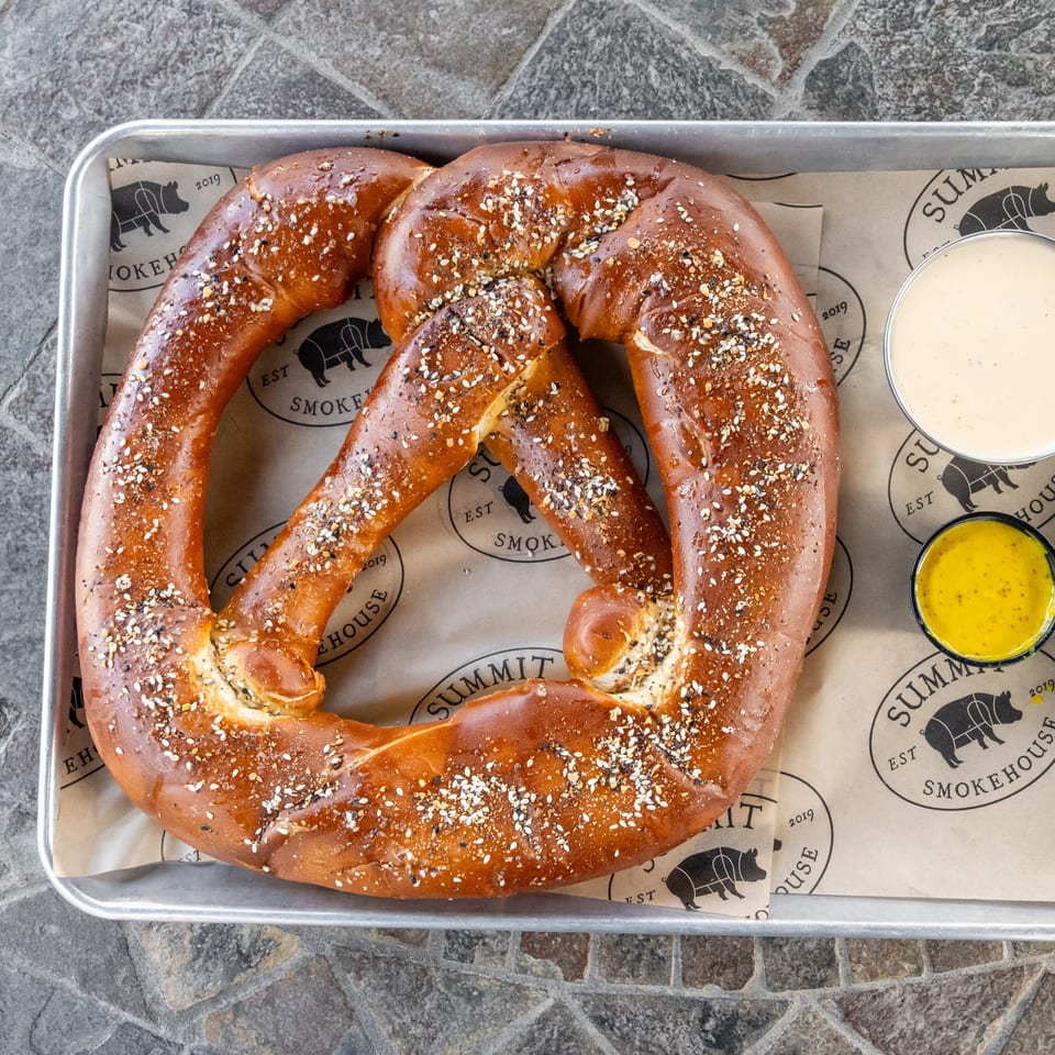 Large seasoned soft pretzel on a metal tray with cups of creamy white and yellow dipping sauces.
