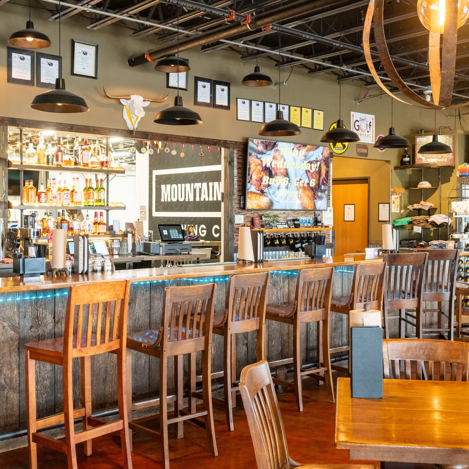 Rustic restaurant bar with wooden chairs, liquor shelves, pendant lighting, sports screens, and a mounted longhorn skull.