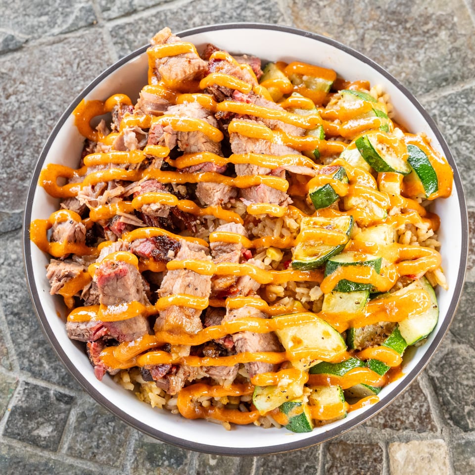 Bowl of sliced grilled steak, rice, and roasted zucchini drizzled with orange sauce on a stone surface.