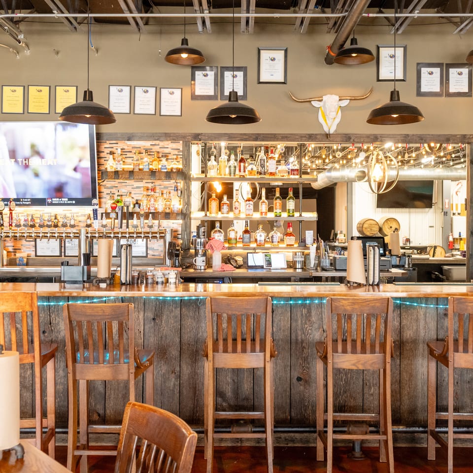 Rustic restaurant bar with liquor shelves, numerous beer taps, televisions, pendant lights, and a long row of wooden chairs.