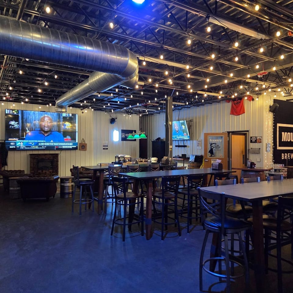 Spacious brewery taproom with high ceilings, exposed ducts, string lights, bar tables, televisions, and a fireplace.