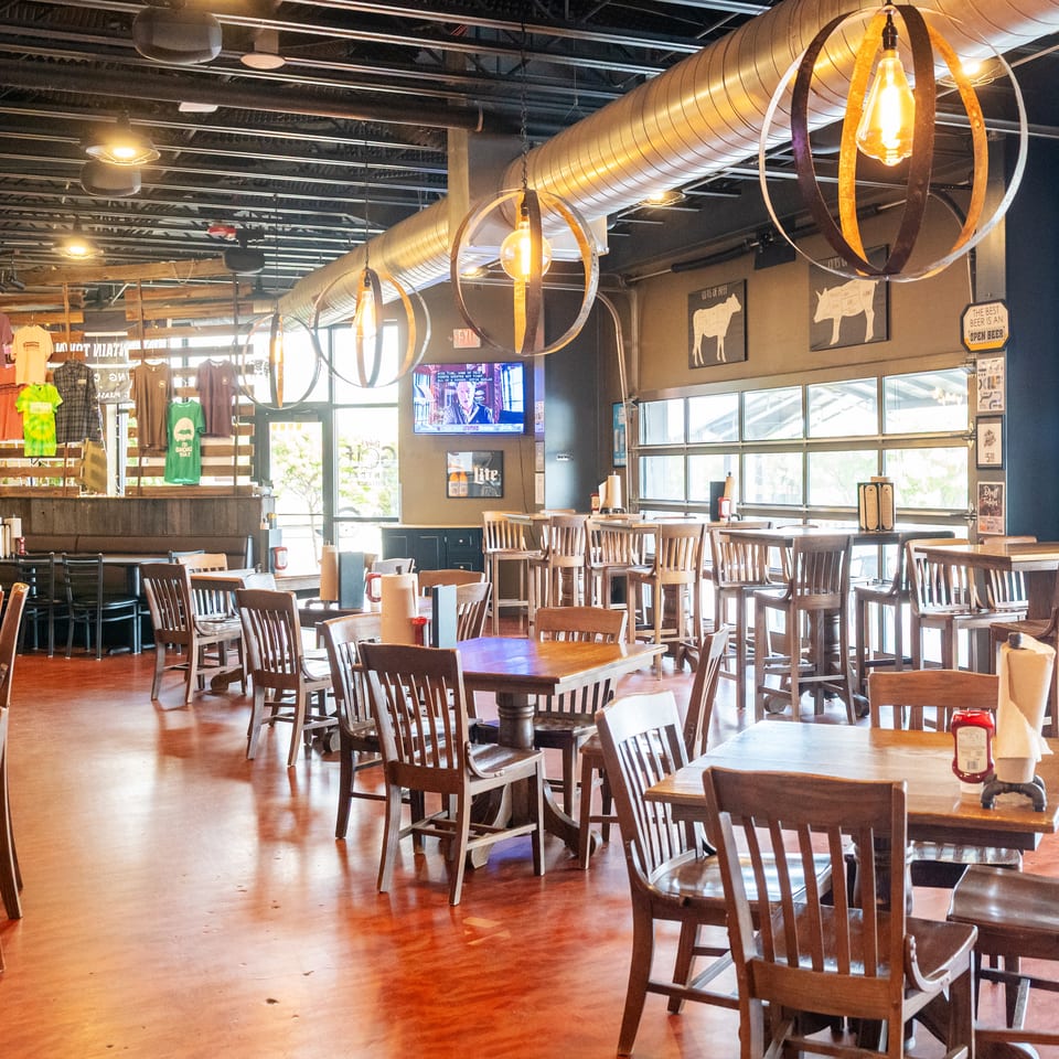 Spacious casual restaurant dining room with wooden tables, chairs, pendant lights, exposed ductwork, merchandise, and beer-themed wall decor.