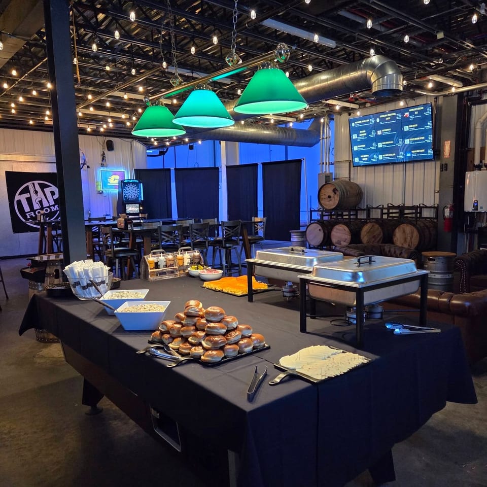 Restaurant interior with buffet table, sandwiches, cheese, green pendant lights, exposed ducts, dining tables, barrels, and digital menu screens.