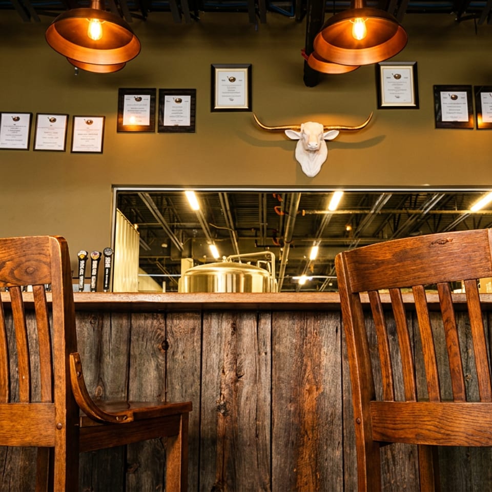 Rustic restaurant bar with two wooden chairs, copper pendant lights, framed certificates, and a longhorn skull mounted above the counter.