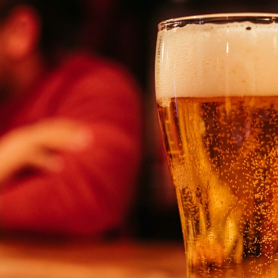 Close-up of a glass of beer with foam, blurred people in the background.