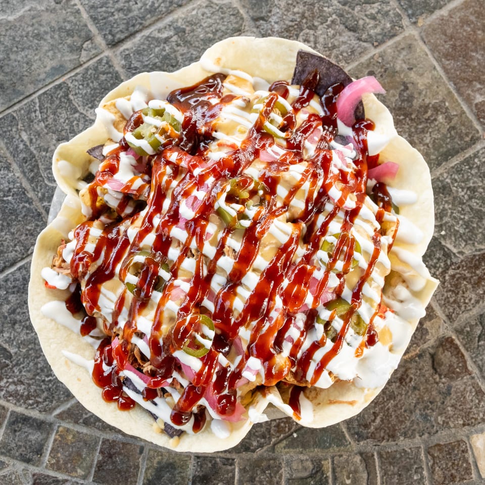 Loaded flatbread topped with shredded meat, pickled onions, jalapeños, creamy sauces, and red sauce on a stone-paved surface.