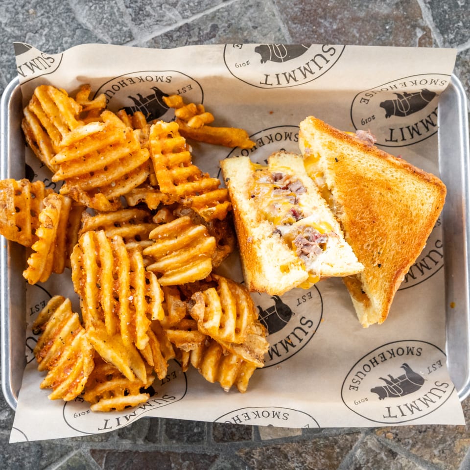 Grilled cheese sandwich with melted cheese and meat served alongside seasoned waffle fries on a paper-lined metal tray.