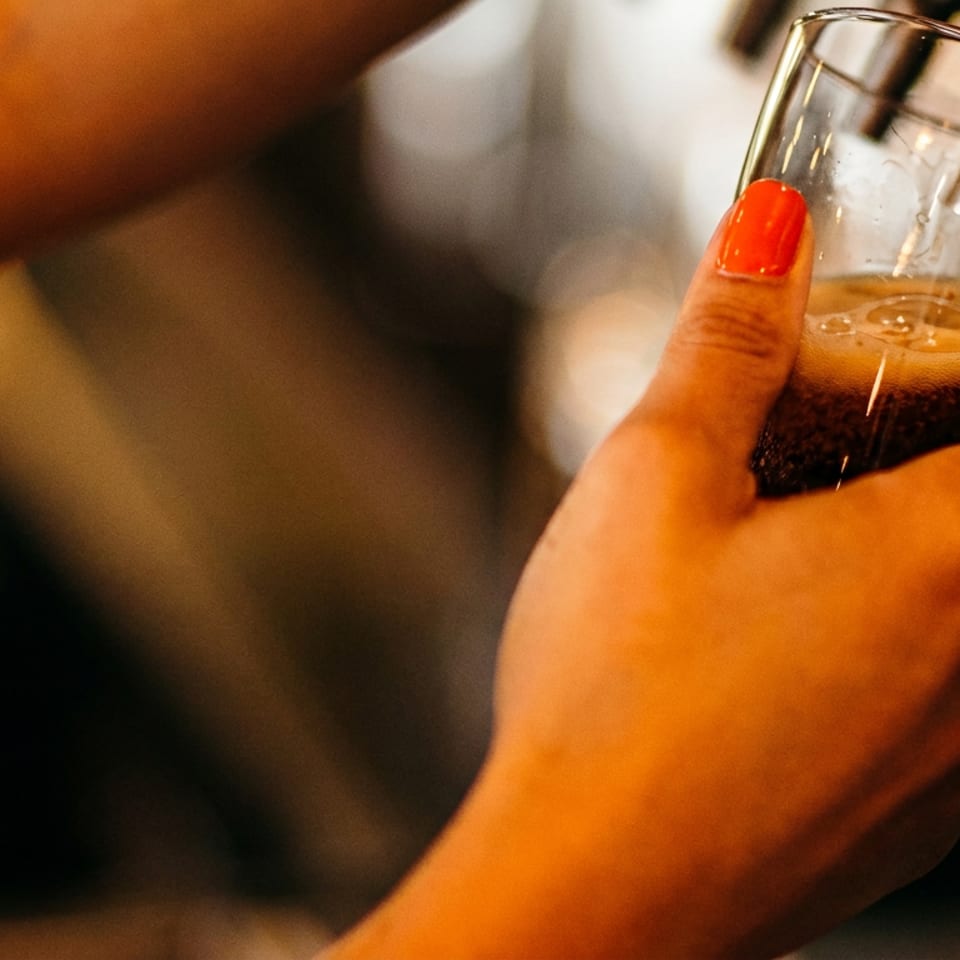A hand with red nail polish holding a glass being filled with a dark beverage from a tap.
