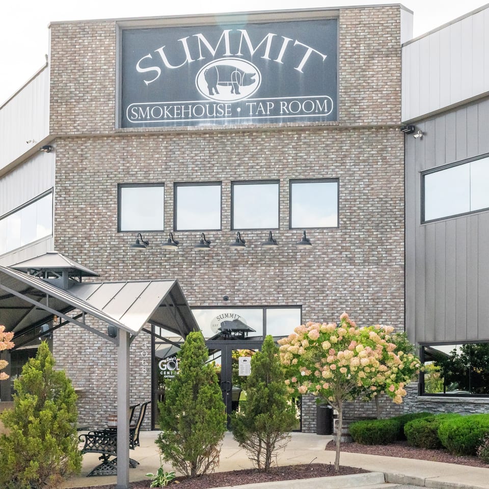 Exterior of Summit Smokehouse Tap Room featuring a brick facade, patio seating, landscaped shrubs, and prominent restaurant signage.